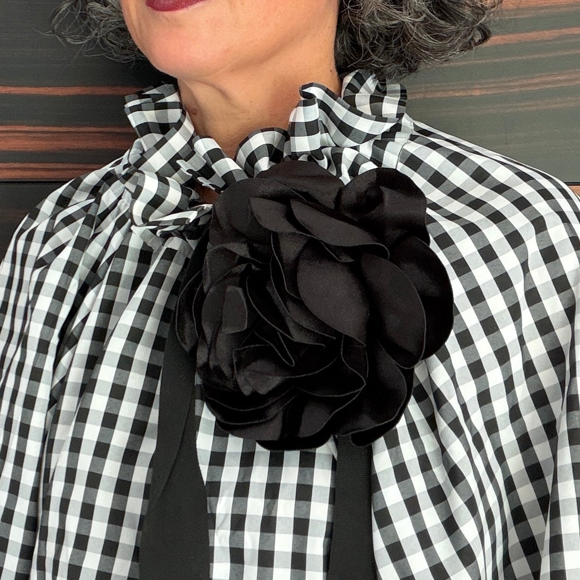 A close-up of a person wearing a black and white gingham blouse with a large black fabric flower accessory at the center of the chest.