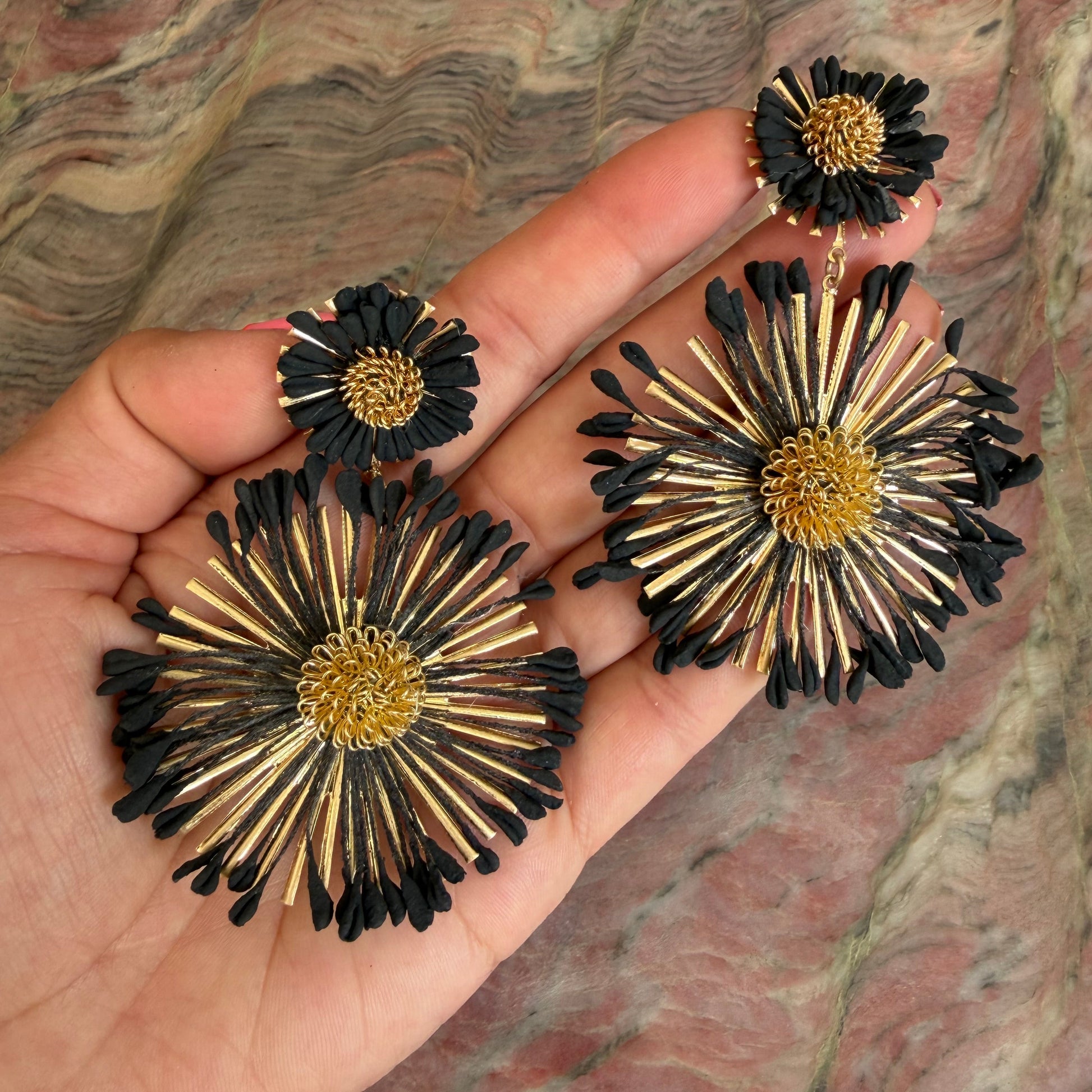 Black and gold flower-shaped earrings held in a hand against a stone background