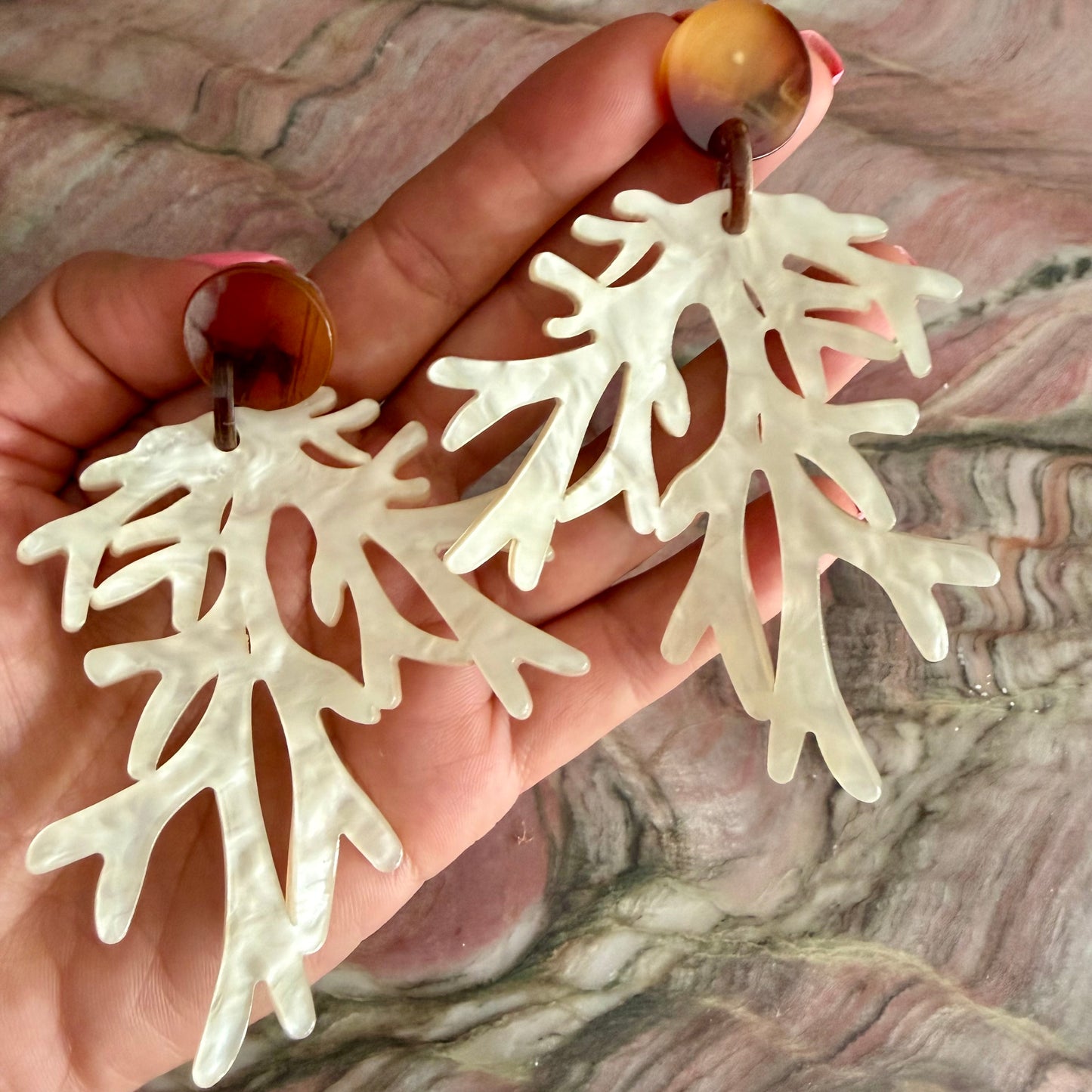 White coral-like earrings held in a hand against a marble background