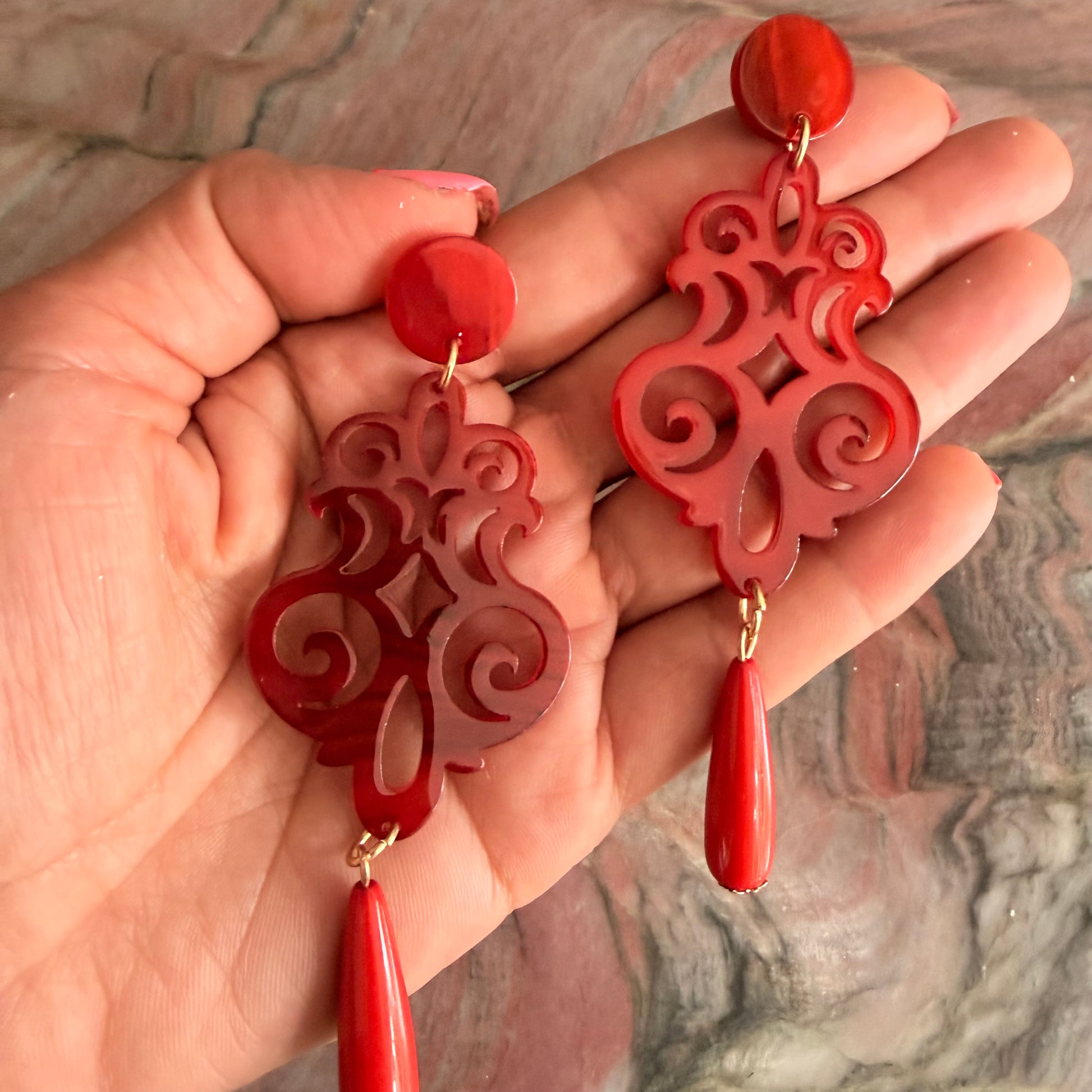 Red earrings with intricate designs held in a hand against a marble background