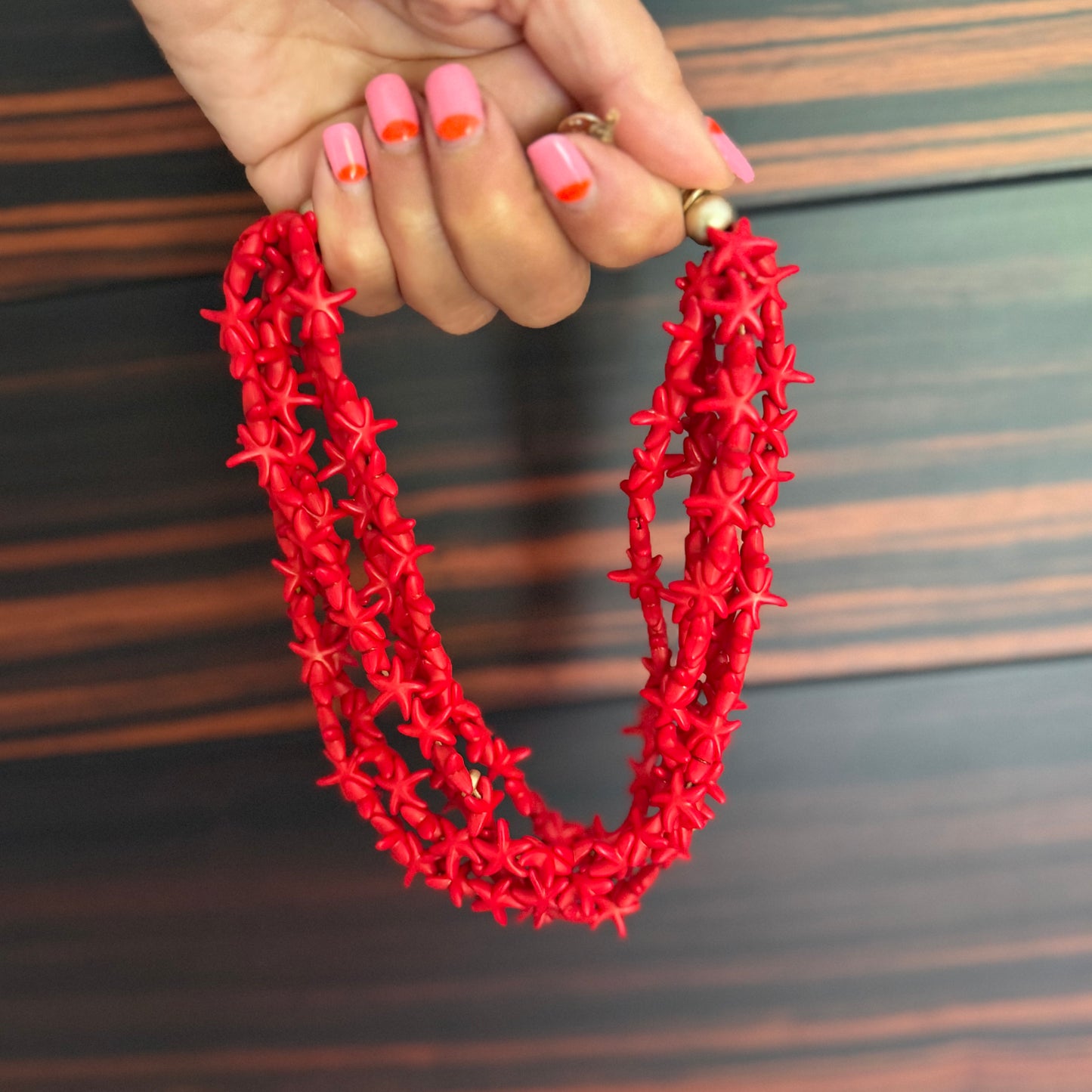 Red necklace held by a hand with pink nail polish on a wooden surface
