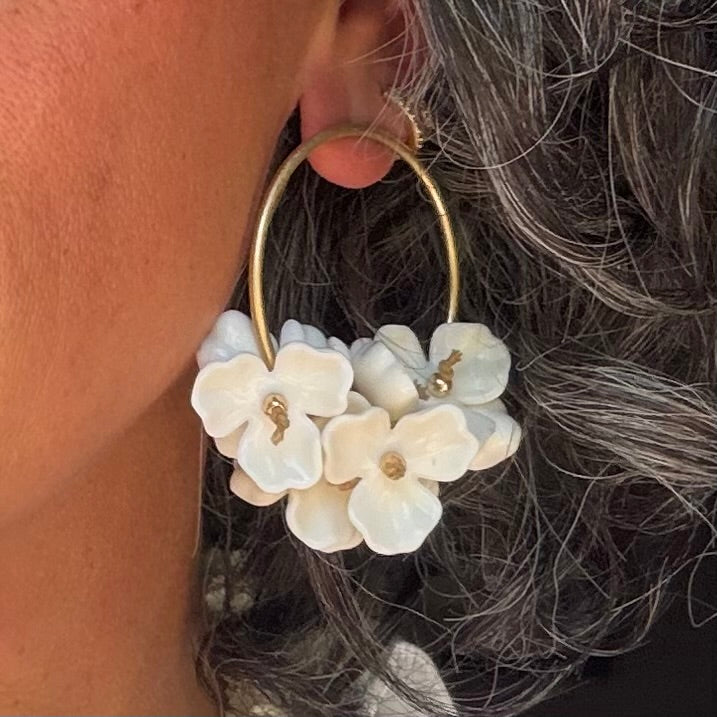 Gold hoop earrings with white floral charms worn by a person with gray hair.